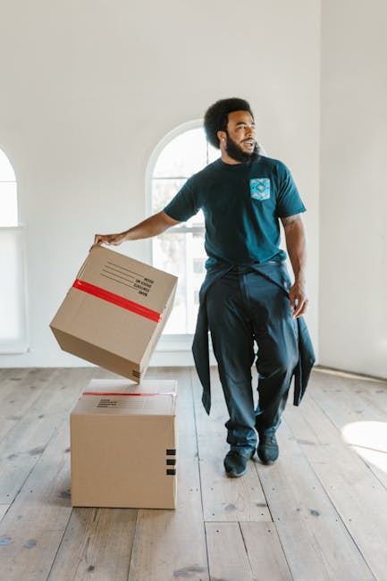 A person wearing light-colored clothing and socks is in the process of packing or unpacking moving boxes indoors, placing a medium-sized cardboard box on top of a neatly stacked pile of similar boxes.