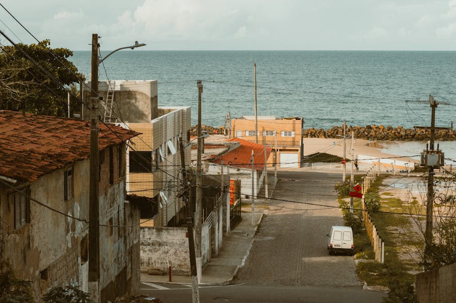 A view of a residential street in a coastal area with a mix of old and new buildings, some with weathered plaster and others with modern facades, situated near the shoreline. Several power poles and overhead electrical wires run along the street, with a streetlamp extending over the pavement. In the foreground, a narrow cobbled road leads down to the water, where a white van is parked close to the edge of the street, near a small fenced area with plants. Beyond the van, the street continues toward the seaside, where large rocks form a breakwater along the shoreline. The scene is illuminated by natural daylight, highlighting the textures of the buildings, pavement, and the calm sea in the background, with a cloudy sky overhead. This setting illustrates a typical coastal neighbourhood suitable for house removals or furniture transport operations, with visual cues relevant to relocating or packing for a move, often managed by companies like Man With a Van Kentish Town.