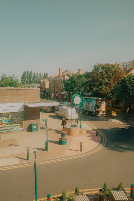 An outdoor street scene in Kentish Town during daylight with a clear sky, showing a parked delivery truck, a white van typical of house removals, and a pedestrian walking near a small shop with a glass storefront. The sidewalk is lined with green bollards and decorative planters, with a couple of traffic cones nearby. Behind the shop, there are residential buildings with pitched roofs and chimneys, and tall trees providing greenery. The street features a curved double yellow line, indicating parking restrictions, and the area appears to be a busy part of the town suitable for furniture transport and house relocation services provided by Man With a Van Kentish Town.