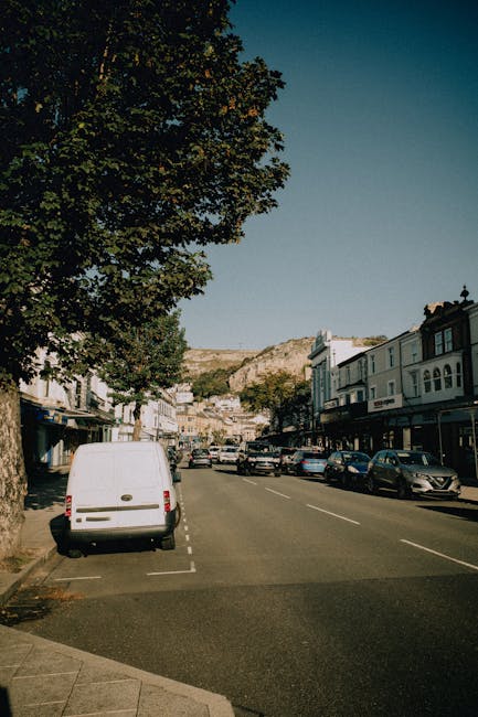 A view of a residential street in a coastal area with a mix of old and new buildings, some with weathered plaster and others with modern facades, situated near the shoreline. Several power poles and overhead electrical wires run along the street, with a streetlamp extending over the pavement. In the foreground, a narrow cobbled road leads down to the water, where a white van is parked close to the edge of the street, near a small fenced area with plants. Beyond the van, the street continues toward the seaside, where large rocks form a breakwater along the shoreline. The scene is illuminated by natural daylight, highlighting the textures of the buildings, pavement, and the calm sea in the background, with a cloudy sky overhead. This setting illustrates a typical coastal neighbourhood suitable for house removals or furniture transport operations, with visual cues relevant to relocating or packing for a move, often managed by companies like Man With a Van Kentish Town.